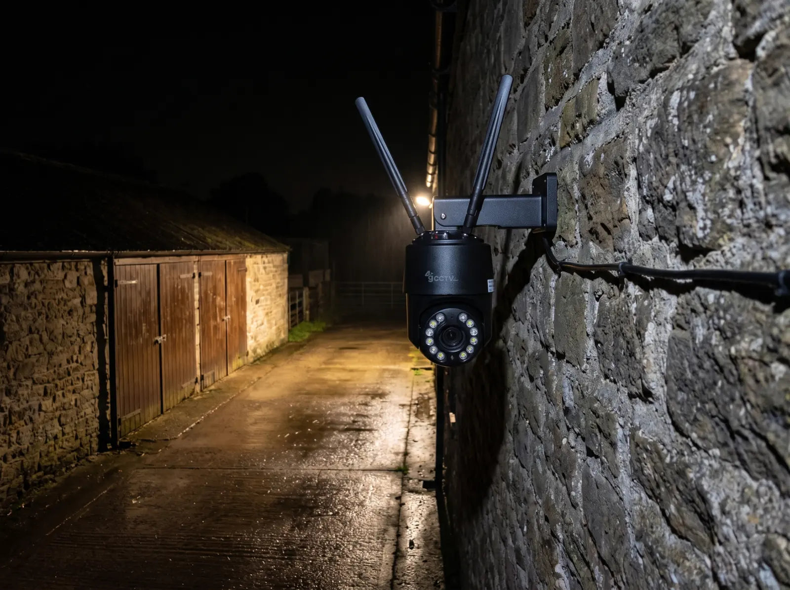 Mini PTZ camera monitoring a wet rural lane beside stone outbuildings at night